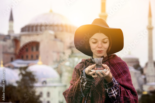A young girl in a burgundy poncho and a hat holds traditional Turkish tea in hands. Hagia Sophia mosque in the rays of the sunset on the background. Travel in Turkey, Istanbul