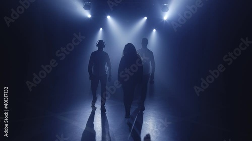 Backlit silhouettes of singer vocalist girl, saxophonist sax, dj man with headphones walking forward in dark nightclub disco for starting performance. Light appears and illuminates musical group band