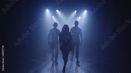Backlit silhouettes of singer vocalist girl, saxophonist sax, dj man walking forward in dark musician nightclub disco for starting concert on stage. Light appears and illuminates musical group band