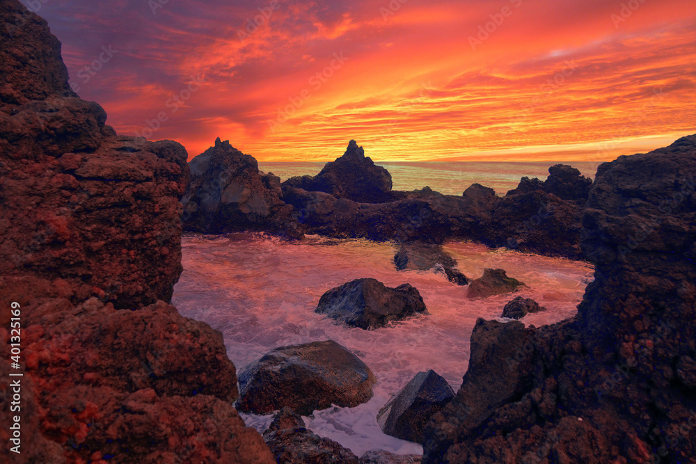 Ocean water fills up a rocky pool area on Pico Island in the Azores Islands during a beautiful sunset with a red, orange, yellow, and magenta sky.
