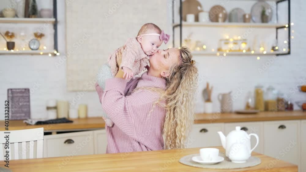 Happy Mother Playing with her Little Daughter in the Kitchen at Home. Mom Lift the Baby up. A Young Woman in Warm Knitted Clothes Hugs and Kisses Enjoying Bonding Cuddling a Infant Girl. Mothers Day Happy Mother Playing with her Little Daughter in the Kitchen at Home. Mom Lift the Baby up. A Young Woman in Warm Knitted Clothes Hugs and Kisses Enjoying Bonding Cuddling a Infant Girl. Mothers Day