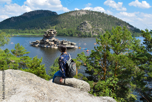 The girl looks at a large stone rock in the middle of a beautiful lake. Stone rocks, mountains, pine trees, tourists on the lake. The girl admires the beautiful views of nature. Burabay, Kazakhstan.