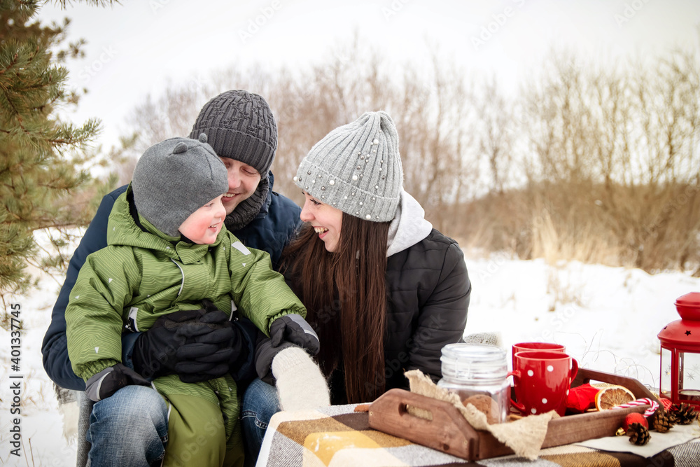 The family in winter in the park on a picnic. Mom, dad and baby sit at the table and look right into the frame. The family in winter in the park on a picnic. Mom, dad and baby sit at the table and look right into the frame.