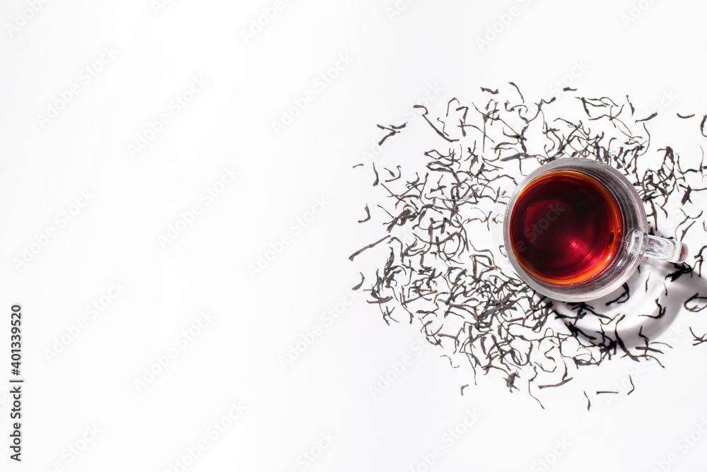 Transparent glass cup of black tea and scattered dry tea leaves. White background, top view copy space