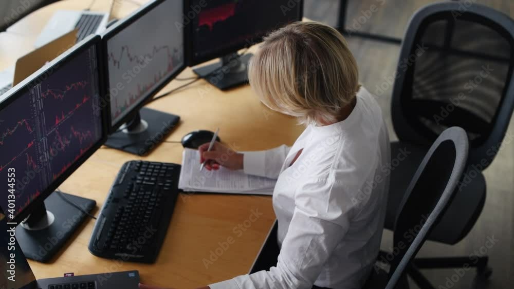 Writing information on the document. Female stockbroker in formal clothes works in the office with financial market and graphs on monitors.
