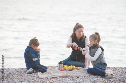 active caucasian family in sleeveless jacket sitting on sea beach drinking hot tea during outdoor leisure vacation travel