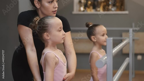 Ballet teacher teaching young dancers. Female ballet teacher helping two little girls in costumes standing with their legs near the barre.