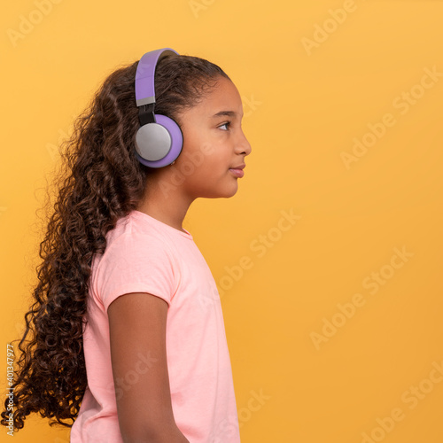 Young dark skinned girl with long curly hair in headphones enjoying music on a yellow background