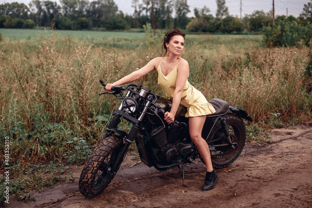 Sexual long-legged attractive female model in a yellow dress sitting on a black motorcycle in the middle of the field and looking to the side against the backdrop of greenery and field. Sexual long-legged attractive female model in a yellow dress sitting on a black motorcycle in the middle of the field and looking to the side against the backdrop of greenery and field.