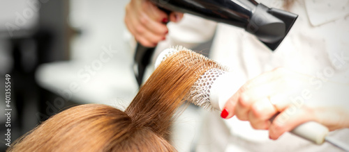 Close up of hairdresser drying long red hair with a hairdryer and round brush in a beauty salon. Selective focus
