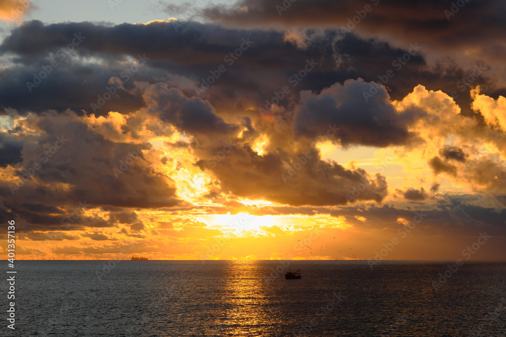 Un tramonto con colori vibranti in un cielo con nuvole nere con stormo di gabbiani che svolazza sopra la barca di un pescatore.