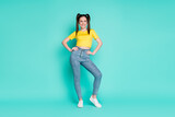 Full size photo of optimistic funky brunette girl stand look empty space wear yellow t-shirt jeans sneakers isolated on cyan background