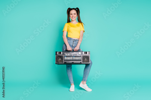 Full size photo of optimistic brunette girl stand hold boombox wear yellow t-shirt jeans sneakers isolated on blue background