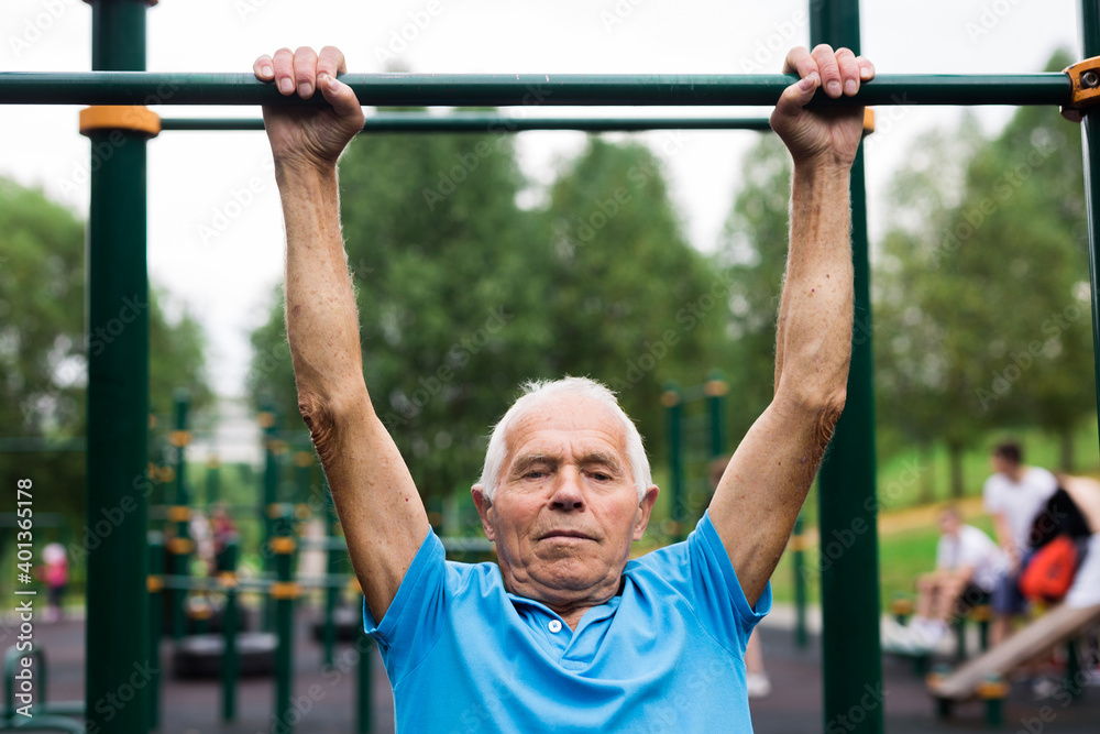 mature cheerful pensioner man doing physical exercises on sports equipped playground