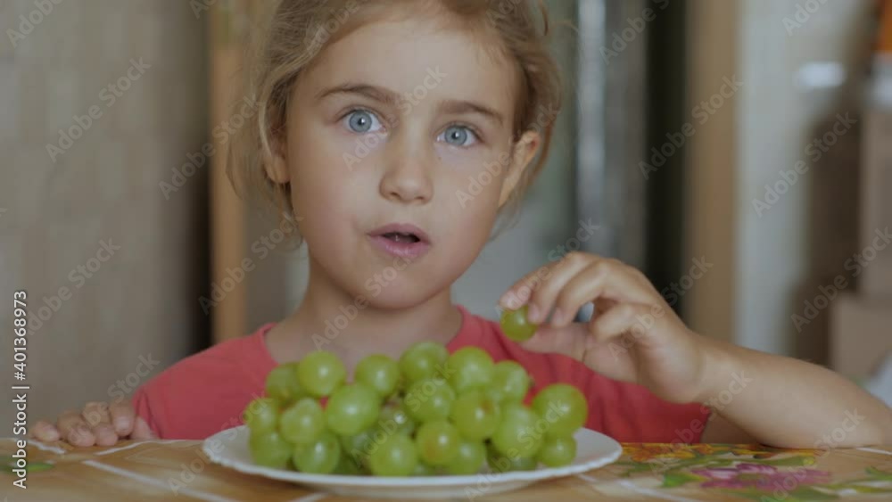 Child Eat Sweet Grape. Little Girl Vegetarian is Eating White Grapes From Plate Sitting Home at Table in Kitchen. Healthy Organic Vegetarian Food. Fruit harvest. Child Eat Sweet Grape. Little Girl Vegetarian is Eating White Grapes From Plate Sitting Home at Table in Kitchen. Healthy Organic Vegetarian Food. Fruit harvest.