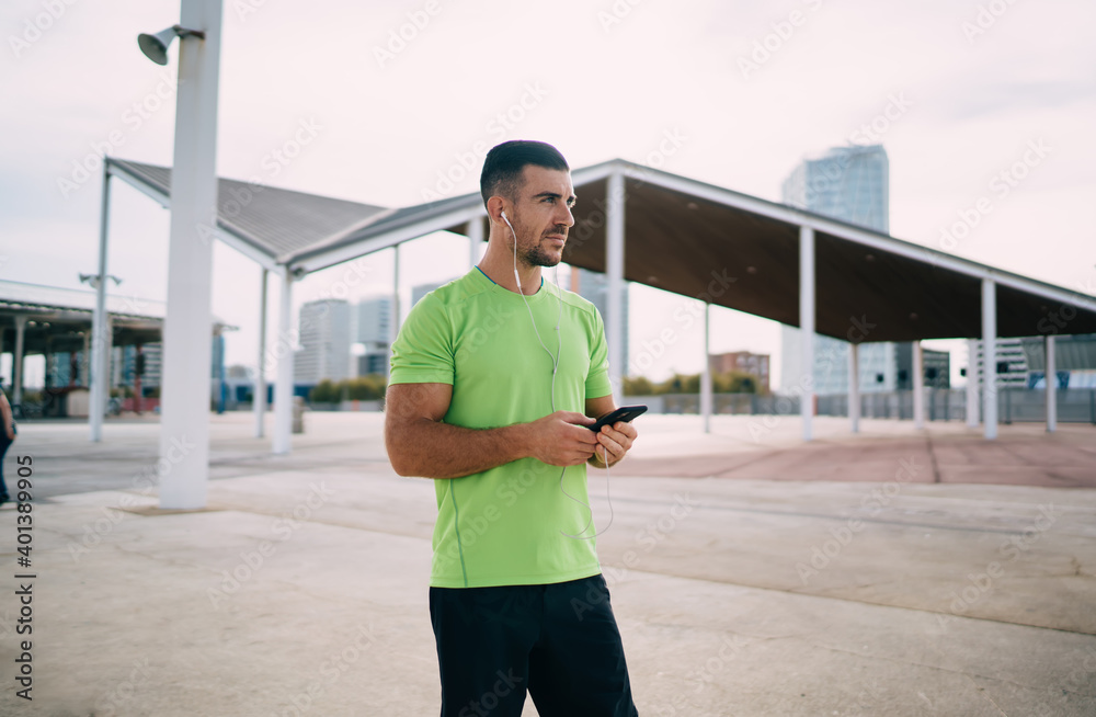 Pondering male athlete in digital headphones using cellular application for listening received audio message with training advices, Caucasian sportsman in earphones thinking about sound record