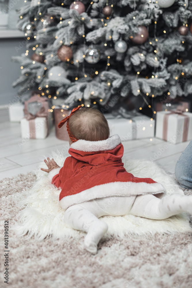 Obraz premium The baby kid at the Christmas tree waiting for Santa. A child looks at a huge Christmas tree and lights. First new year.