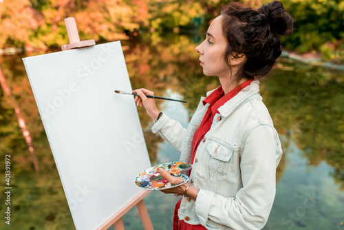 woman artist summer forest by river  draws picture  beginning of landscape creation of creativity by lake pond. White canvas painting with tassel and color palette. Denim jacket  girl s hair in bun.