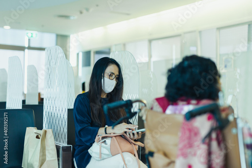 Woman in protective mask using cell phone sitting in partition with her senior mother in hospital.