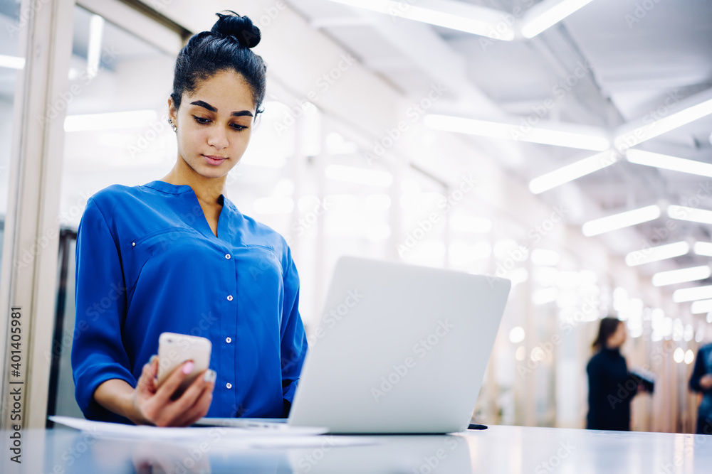 Serious ethnic businesswoman looking at smartphone while working on laptop Serious ethnic businesswoman looking at smartphone while working on laptop