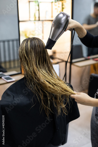 Happy young woman and hairdresser with fan making hot styling at hair salon