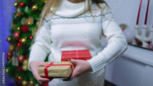 A girl near a Christmas tree and a fireplace with gifts in her hands chooses one to show it to in focus. New Year's gifts for Christmas