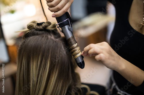 Young woman hairdresser making curls at blond hair with curling irons at luxury barber salon. Stylist and beauty