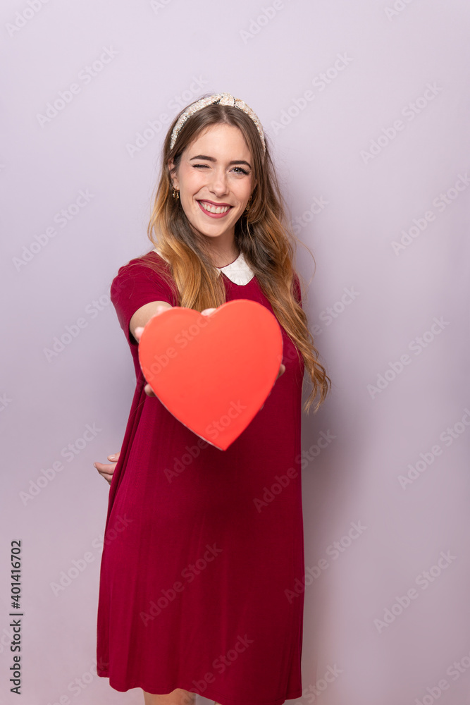 Cute young woman holding a red heart shaped valentiens gift box to the camera for copy text and wink an eye Cute young woman holding a red heart shaped valentiens gift box to the camera for copy text and wink an eye