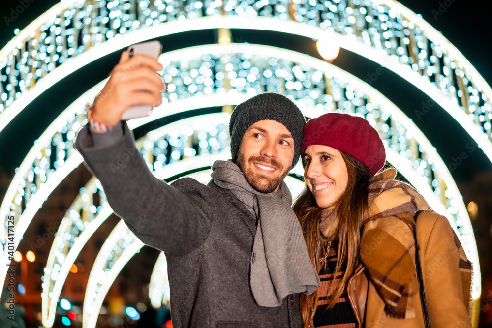 Winter portrait of a Caucasian couple enjoying the Christmas lights of the city taking a selfie with the mobile for social networks, lifestyle