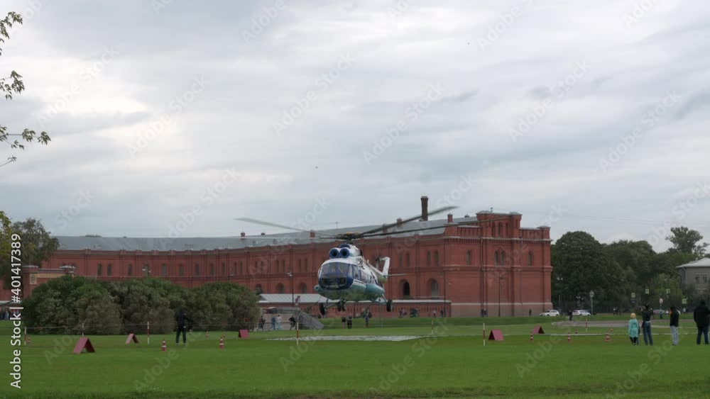 A helicopter with large blades takes off over the helipad. The helicopter took off over a green lawn and people in the city A helicopter with large blades takes off over the helipad. The helicopter took off over a green lawn and people in the city