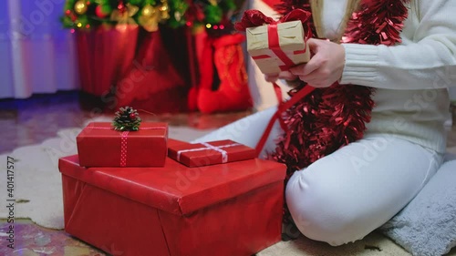 A girl in a white sweater sits at home by a Christmas tree and ties a ribbon on a red box with a New Year's gift