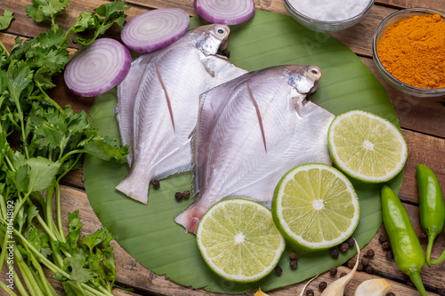Fresh white pomfret fish along with onion, garlic, coriander leaves, lemon and spices on wood background.