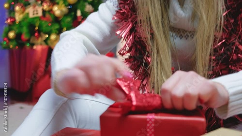 A cheerful girl in a white sweater sits at home by the Christmas tree and wraps a box with a gift. moving the camera forward