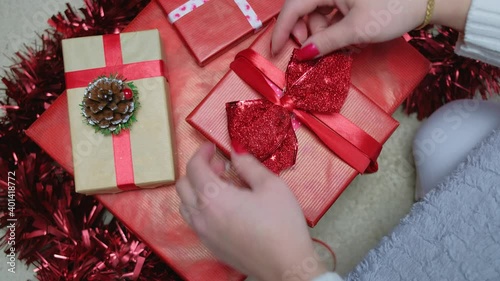 A girl sits at home by a Christmas tree and ties a ribbon on a red box with a New Year's gift, the camera moves from top to bottom