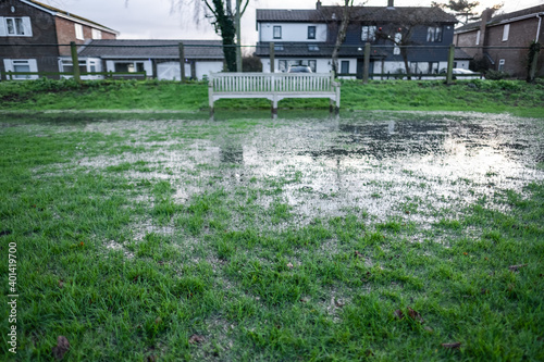 Shallow focus of grass seen on a public park having been subjected to heaving storms. The area is now waterlogged and a distant park bench is visible.