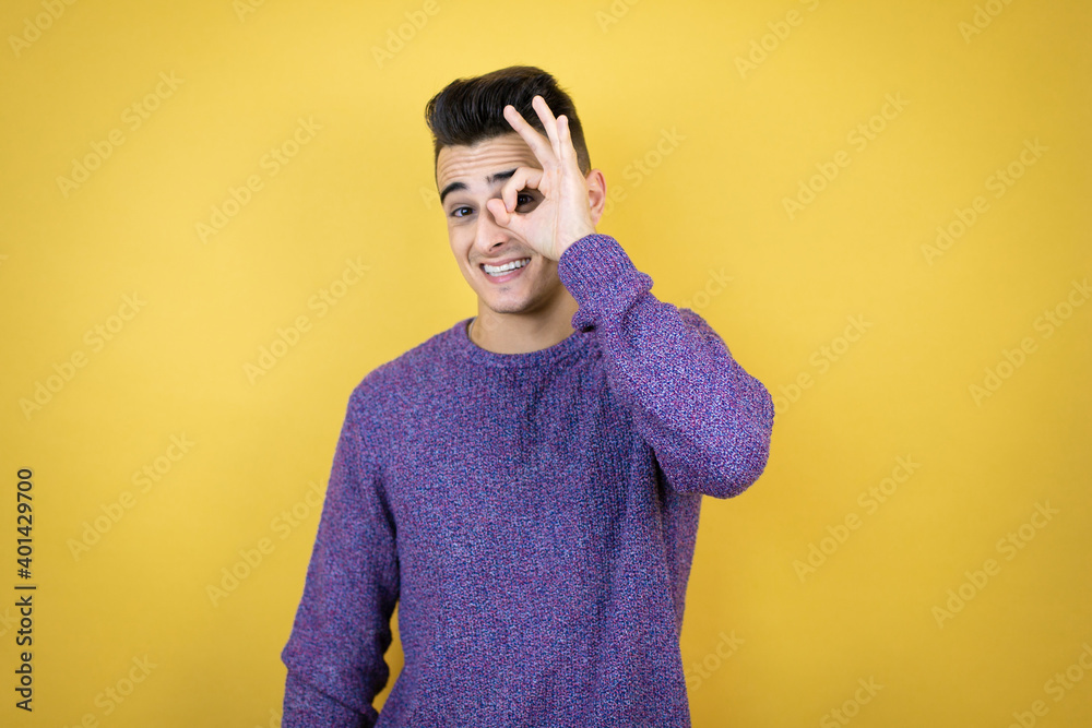 Young caucasian man over isolated yellow background doing ok gesture shocked with smiling face, eye looking through fingers