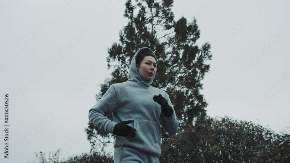 Young woman in sportswear and gloves jogging outside near trees and bushes, sky on background. Low angle dolly shot of fit female training in the morning. Concept of sport Young woman in sportswear and gloves jogging outside near trees and bushes, sky on background. Low angle dolly shot of fit female training in the morning. Concept of sport