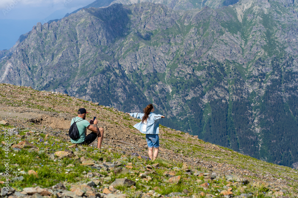 Rear view of man photographing woman on smartphone in mountains. Male takes pictures his female on background of mountain landscape in sunny weather.