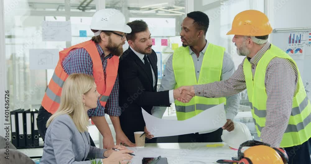 Attractive satisfied experienced bearded customer handshaking with main man of contractors of building company during meeting where they discussing building project with team of contractors in office Attractive satisfied experienced bearded customer handshaking with main man of contractors of building company during meeting where they discussing building project with team of contractors in office