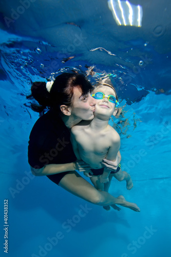 A beautiful little boy and his mother are swimming together under the water. She kisses him gently on the cheek. Active happy child. Family sports lessons. Healthy lifestyle
