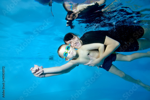 A beautiful boy and a female coach swim under the water with their arms outstretched. They smile and look at the camera. Active happy child. Swimming lessons for small children. Healthy lifestyle