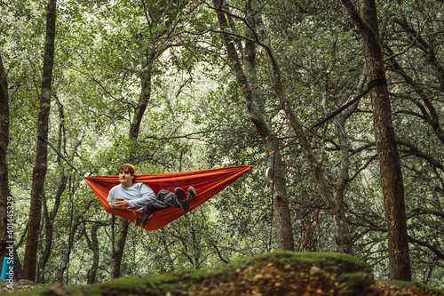 Hammock on trees in the forest, man lying in a hammock in the middle of a dark forest, orange hammock, man wears teal sweater and orange hat