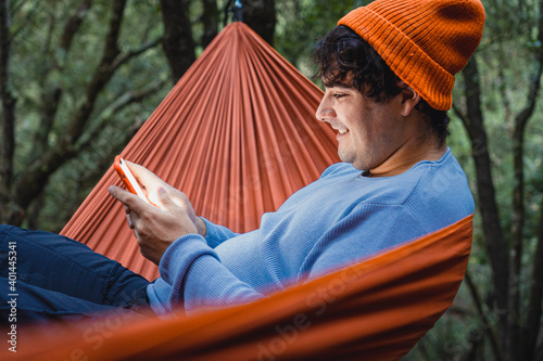 Hammock on trees in the forest ,portrait of man using mobile from inside a hammock in the forest, wears teal sweater and orange hat