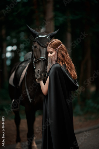 Young woman in hat and black cloak with a horse in the forest