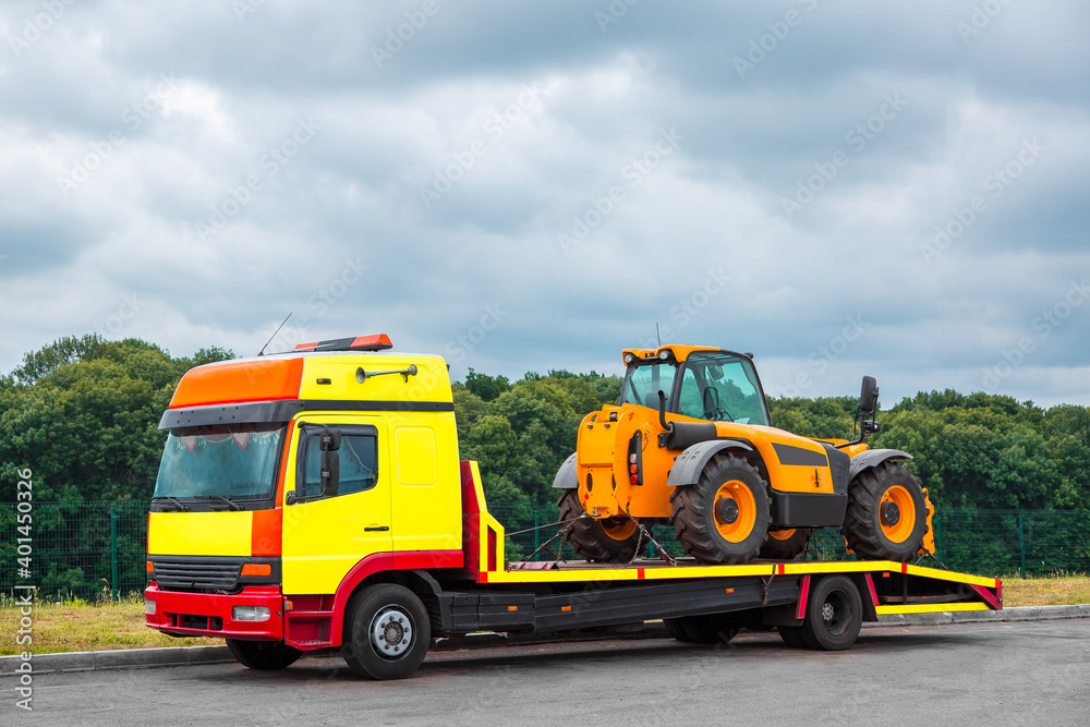 The tow truck on the platform transports the new tractor, equipment for technical and agricultural works stands on an asphalt road near the curb, in the background a forest in cloudy weather, nobody.