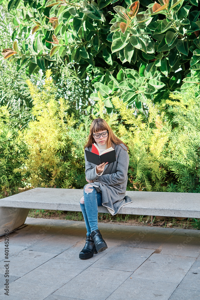 Blue-eyed Caucasian blonde young woman with glasses reading a book in the park