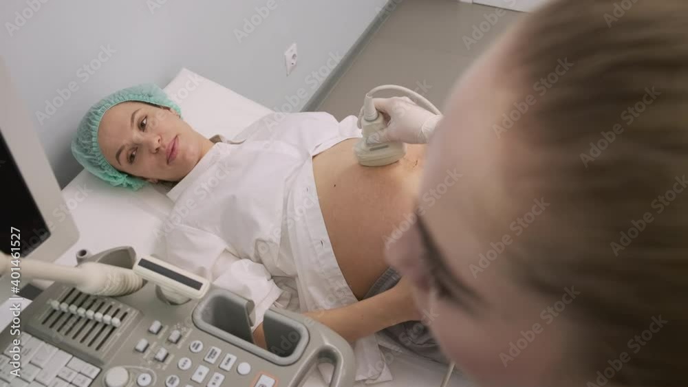 Close-up. A young girl doctor in a white coat conducts ultrasound diagnostics to a young pregnant patient in a clinic office Close-up. A young girl doctor in a white coat conducts ultrasound diagnostics to a young pregnant patient in a clinic office