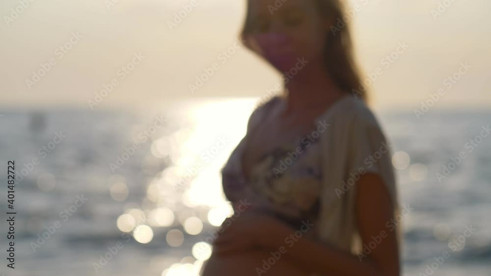 beautiful unrecognizable defocused pregnant woman at sea at sunrise, sparkling water close up of pregnant belly, happy maternity concept