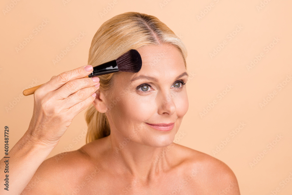 Close-up portrait of lovely middle aged lady applying natural powder correction tone isolated over beige pastel color background