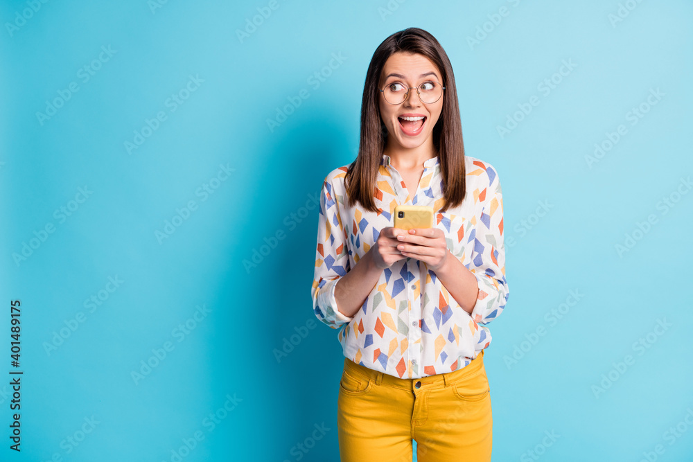 Photo of cute girl hold smartphone look side empty space wear glasses geometry print shirt yellow pants isolated blue color background Photo of cute girl hold smartphone look side empty space wear glasses geometry print shirt yellow pants isolated blue color background